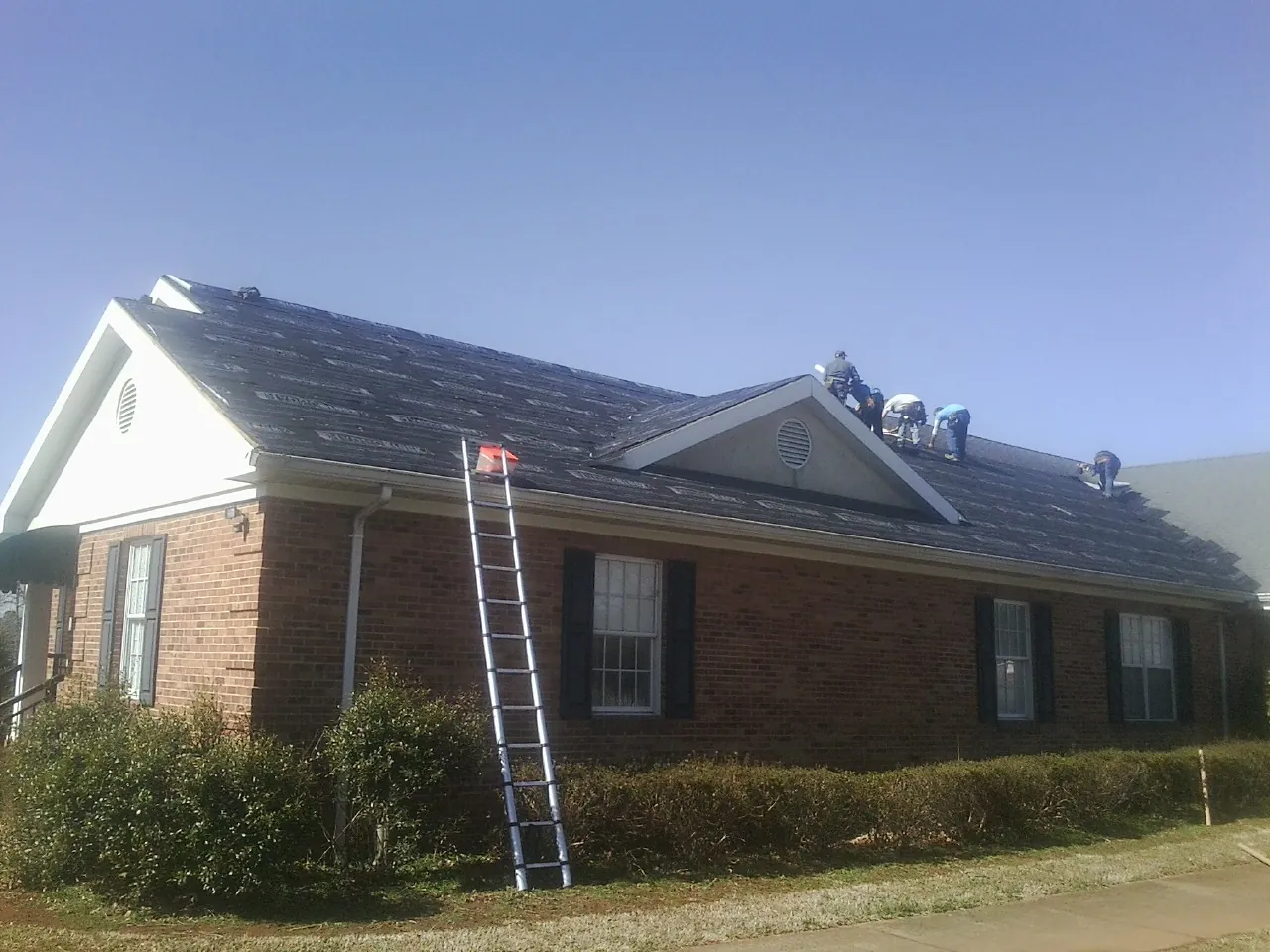 Crew tearing shingles off a brick ranch roof with a ladder set — Metal Roof Repair work by our crew in Lakota