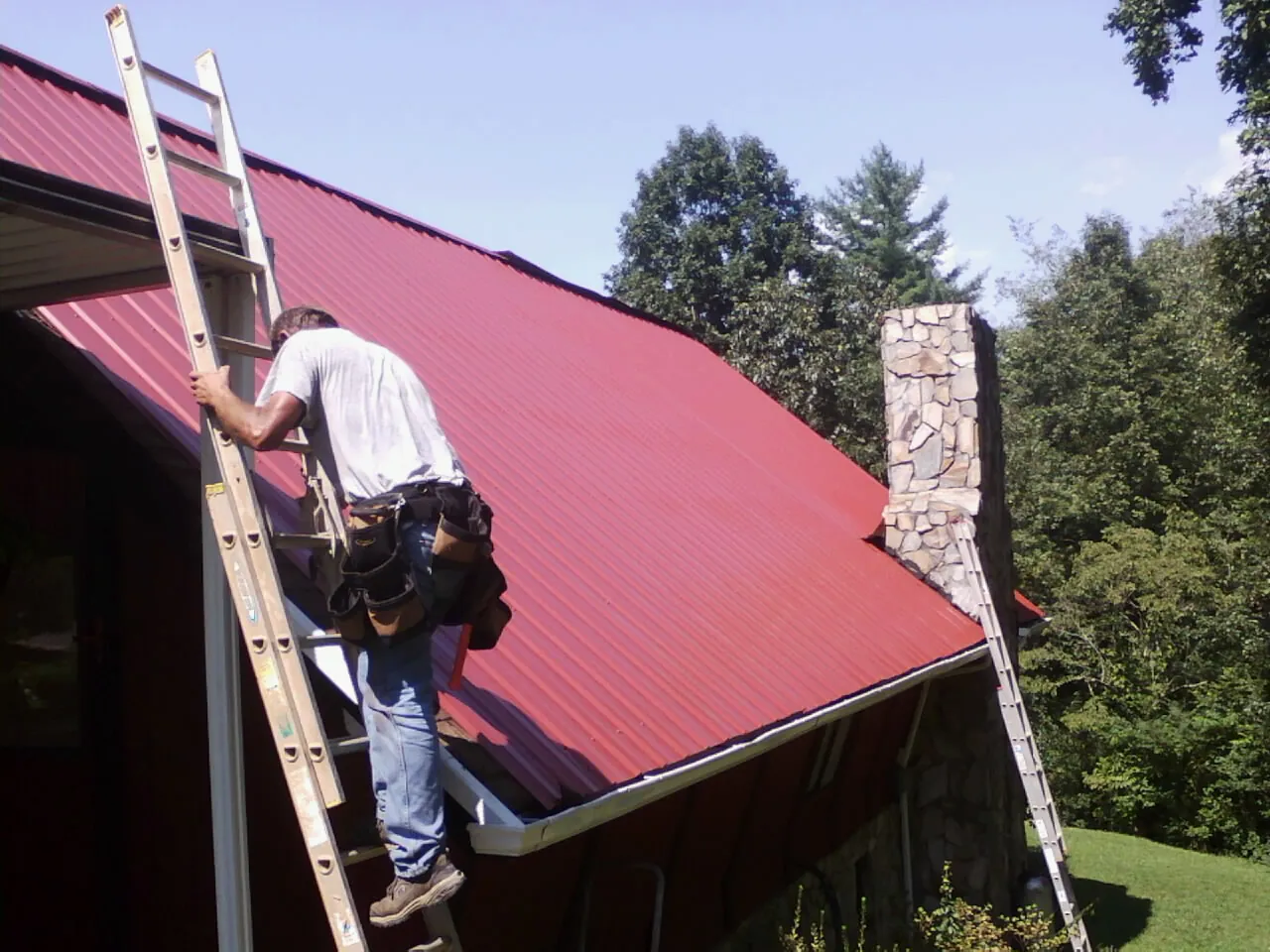 Roofer on a ladder beside a red metal roof and stone chimney — Soffit & Fascia Repair by Roofing Brothers in Lakota